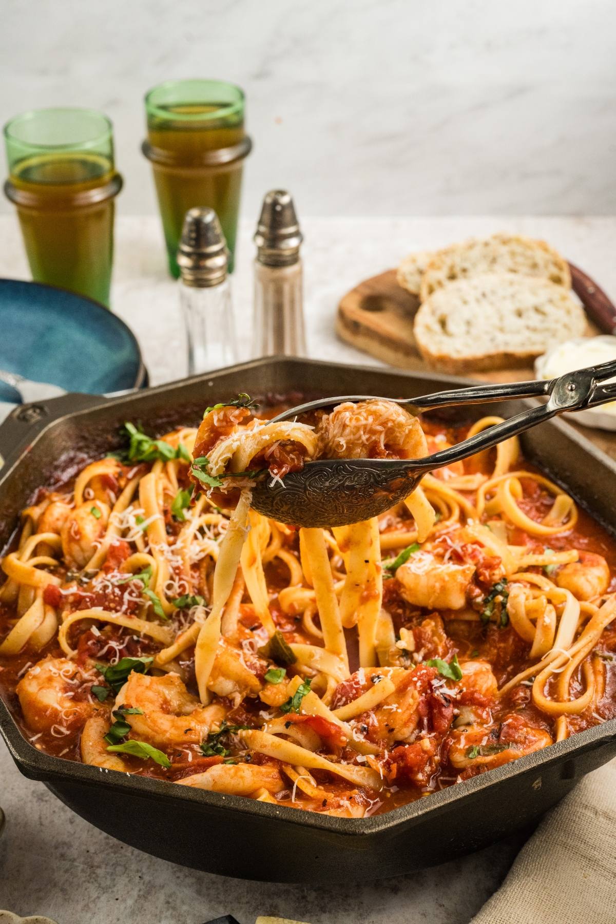 Tongs lifting Shrimp fra diavolo  pasta from a skillet of fettuccine with tomato sauce, shrimp, and cheese, with bread in background.