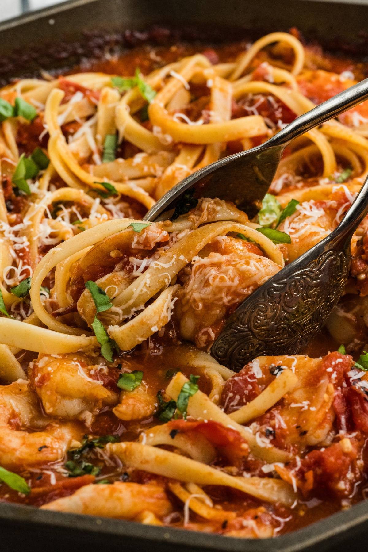 Pasta with tomato sauce, chicken, and basil being served with tongs in a black pan, topped with grated cheese.