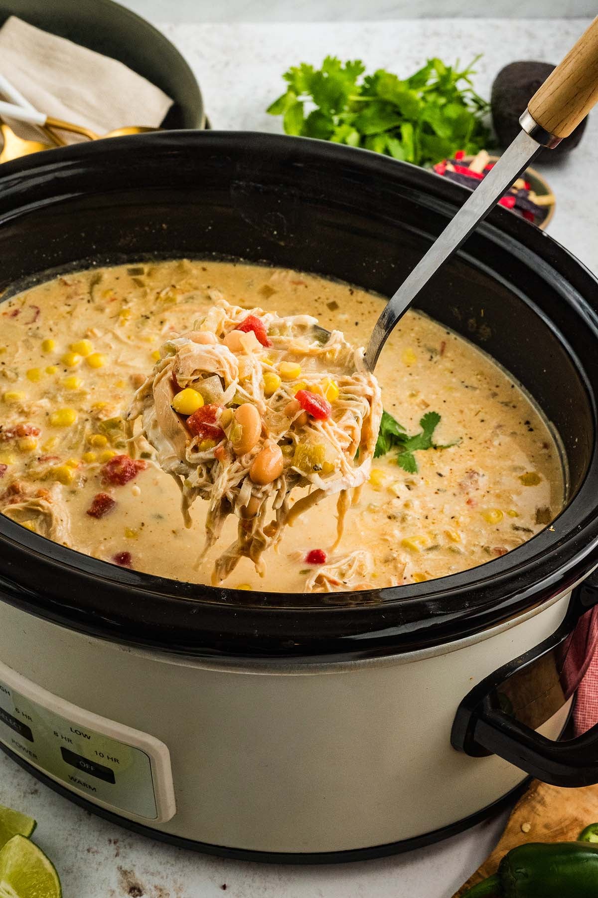 A ladle scoops creamy Slow Cooker Green Chile Chicken Soup with beans, corn, and tomatoes from a slow cooker.