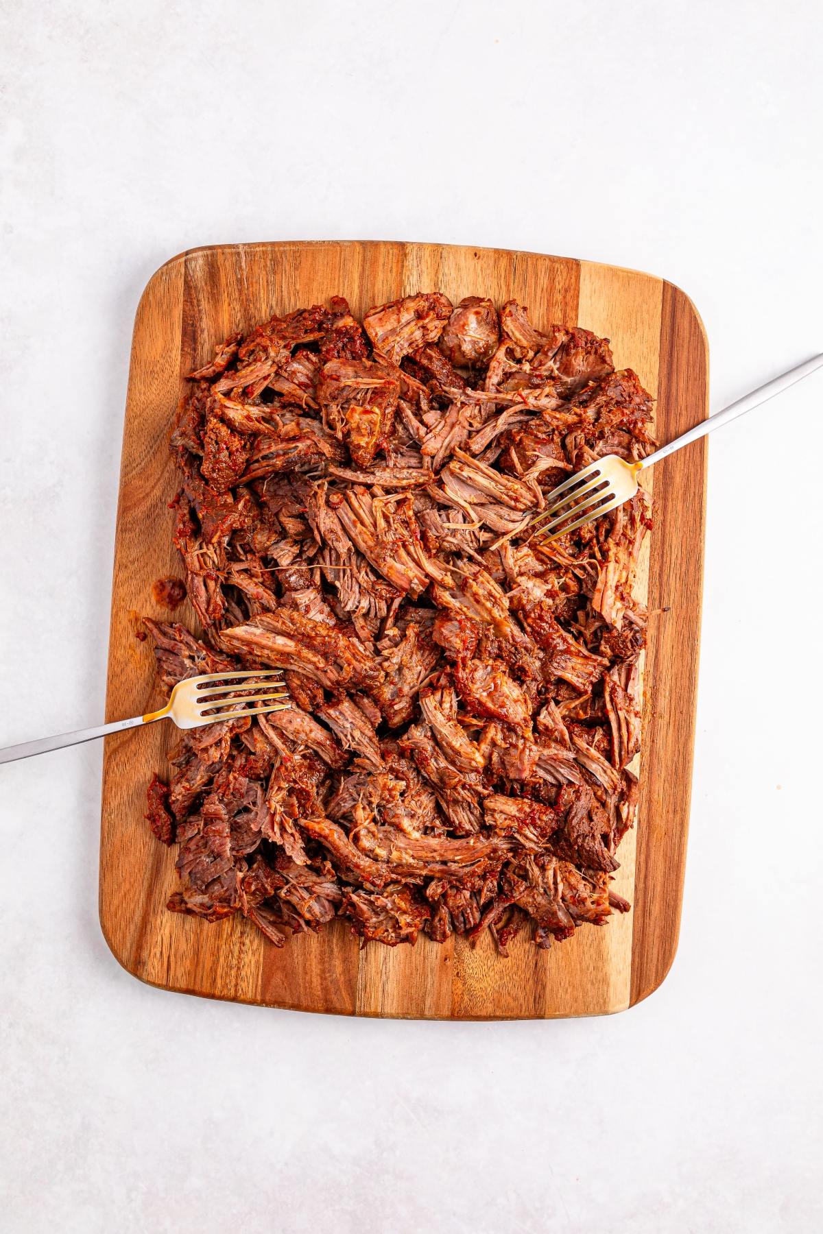 Shredded beef on a wooden cutting board with two forks pulling the meat apart.