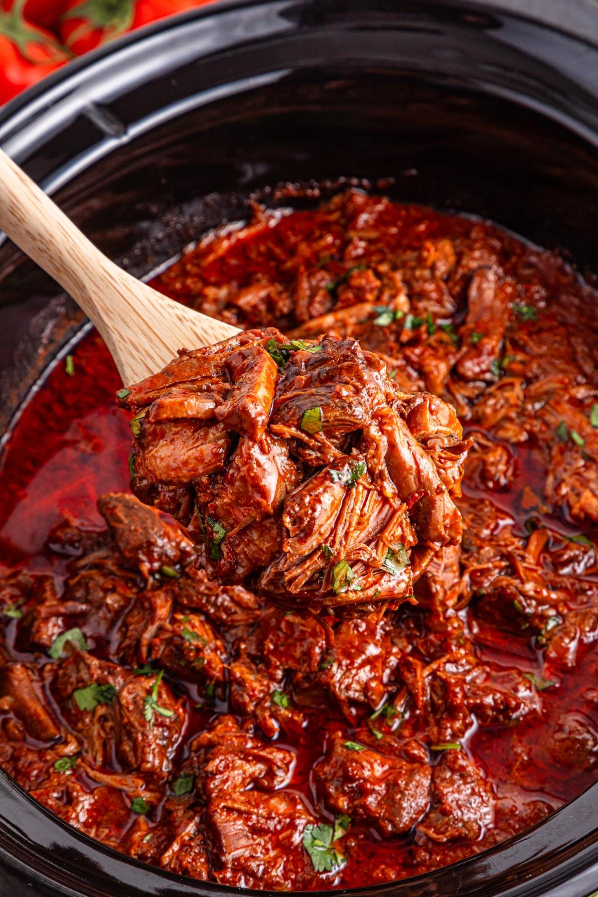 A wooden spoon lifts shredded, saucy meat from a slow cooker filled with rich, red stew.