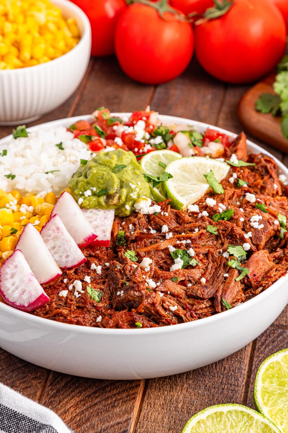 A slow cooker birria with shredded beef, rice, corn, guacamole, radishes, pico de gallo, and lime slices.