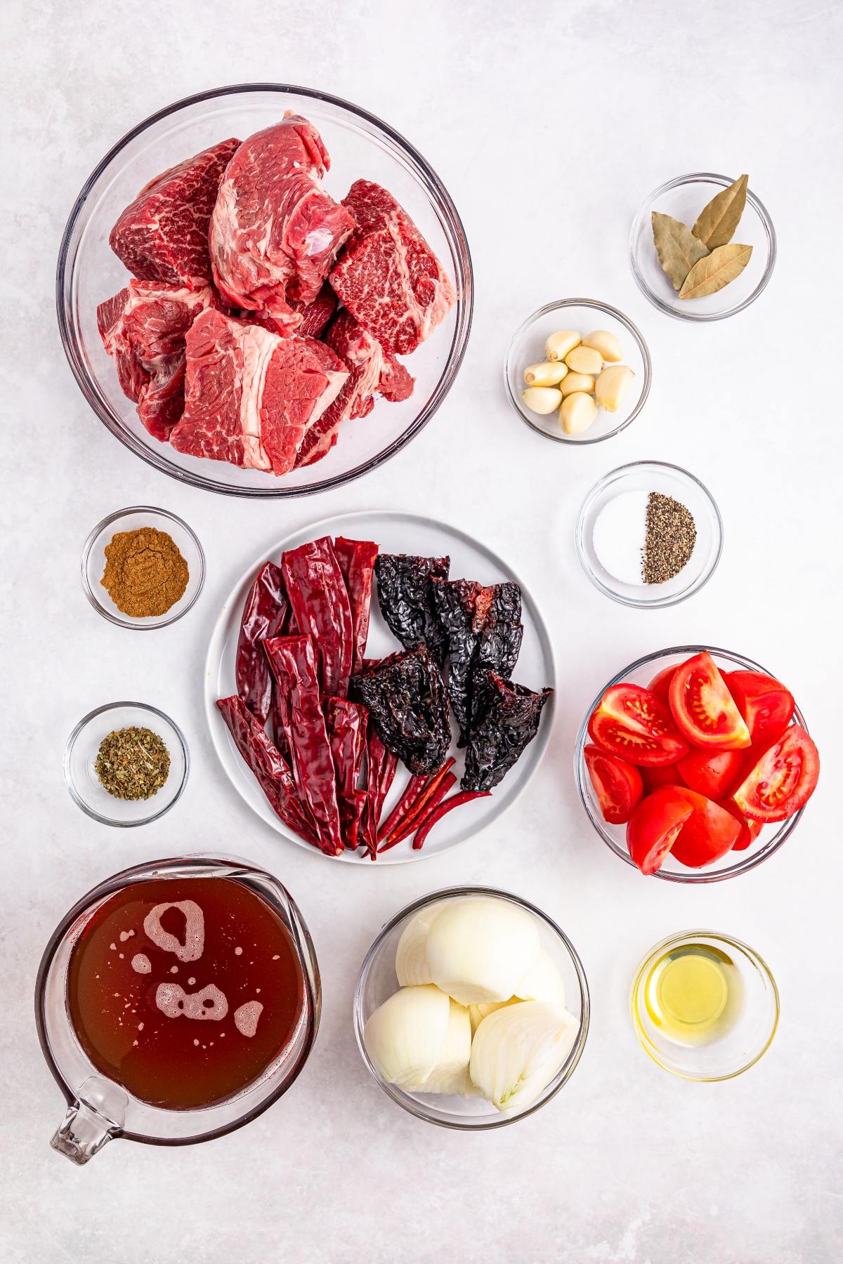 Bowls of raw beef, dried chilies, tomatoes, onions, garlic, spices, bay leaves, broth, and oil on a white surface.
