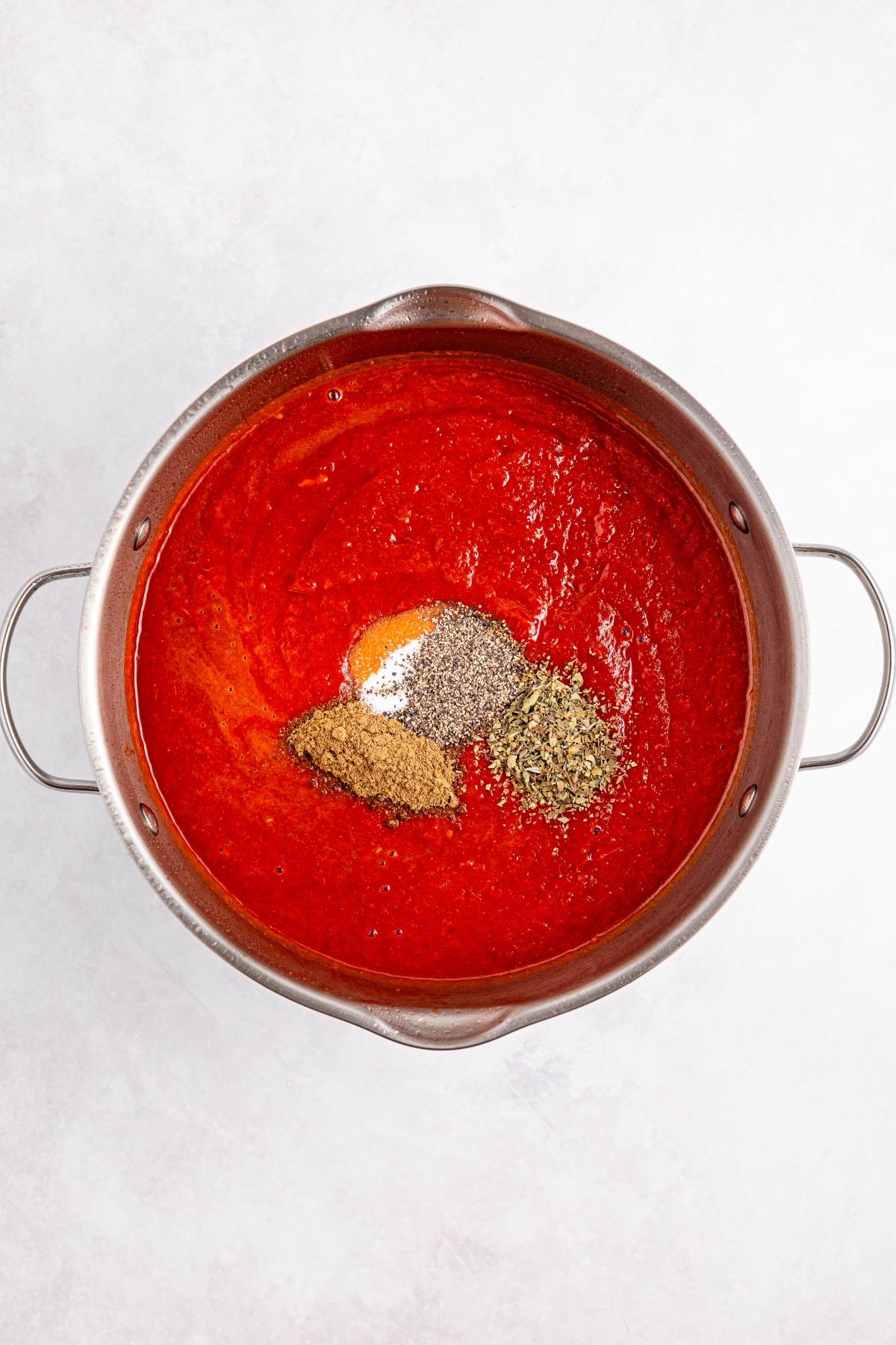 A pot of tomato sauce with piles of spices on top, viewed from above on a light surface.