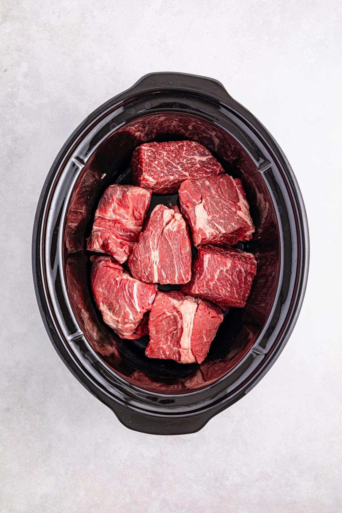Chunks of raw beef placed inside a black slow cooker on a light, textured surface.