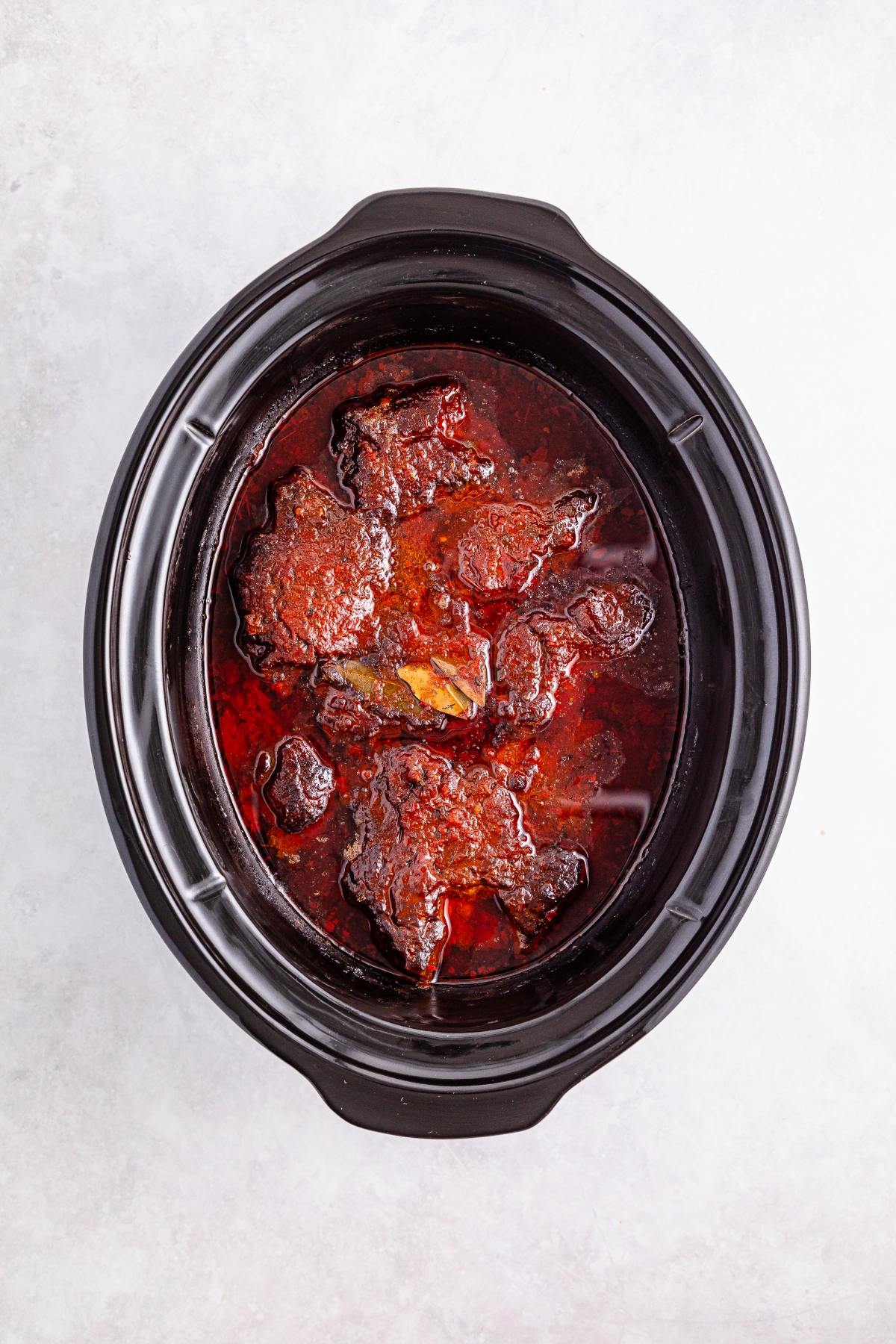 Beef stew with rich red sauce in a black slow cooker, viewed from above on a light background.