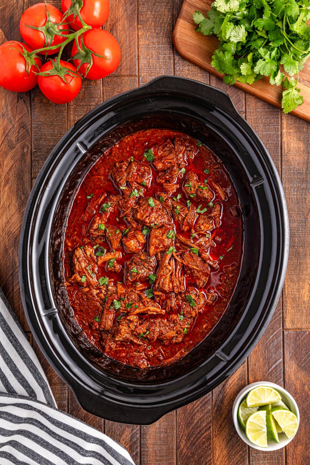 Slow cooker birria filled with shredded beef in red sauce, surrounded by tomatoes, cilantro, lime, and a striped towel.