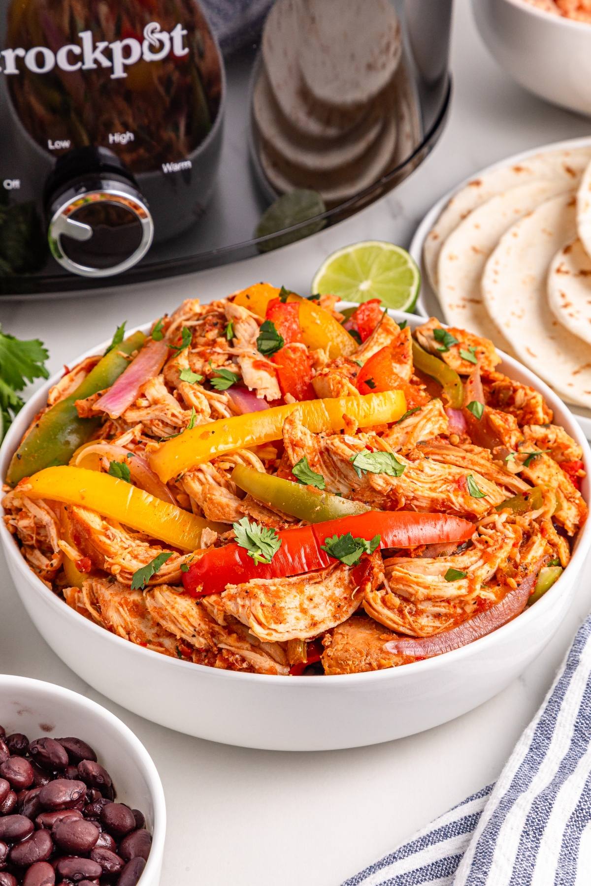 A bowl of shredded slow cooker chicken fajitas with bell peppers, served near tortillas, black beans, and a slow cooker.