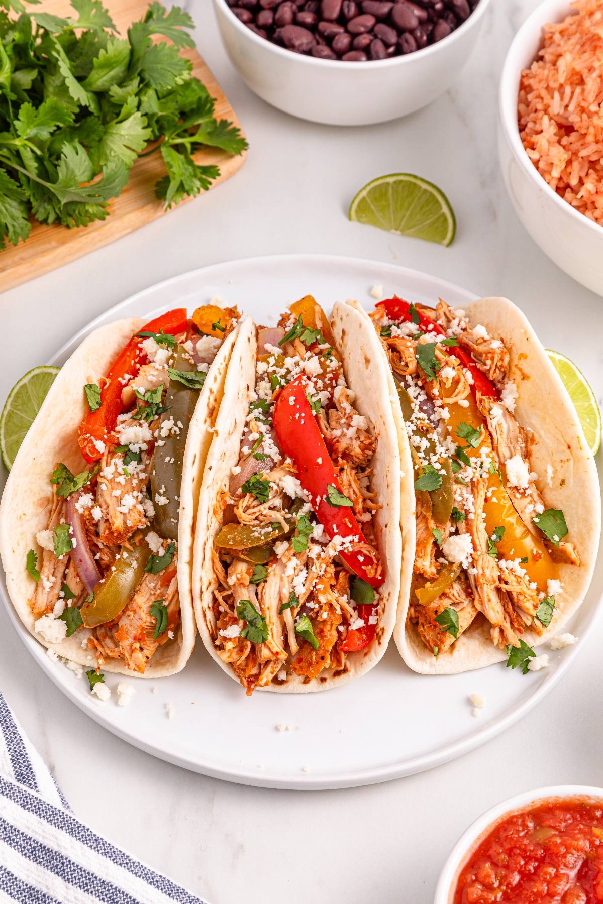 Three slow cooker chicken fajita tacos with colorful peppers, cheese, and cilantro on a plate, surrounded by rice and beans.