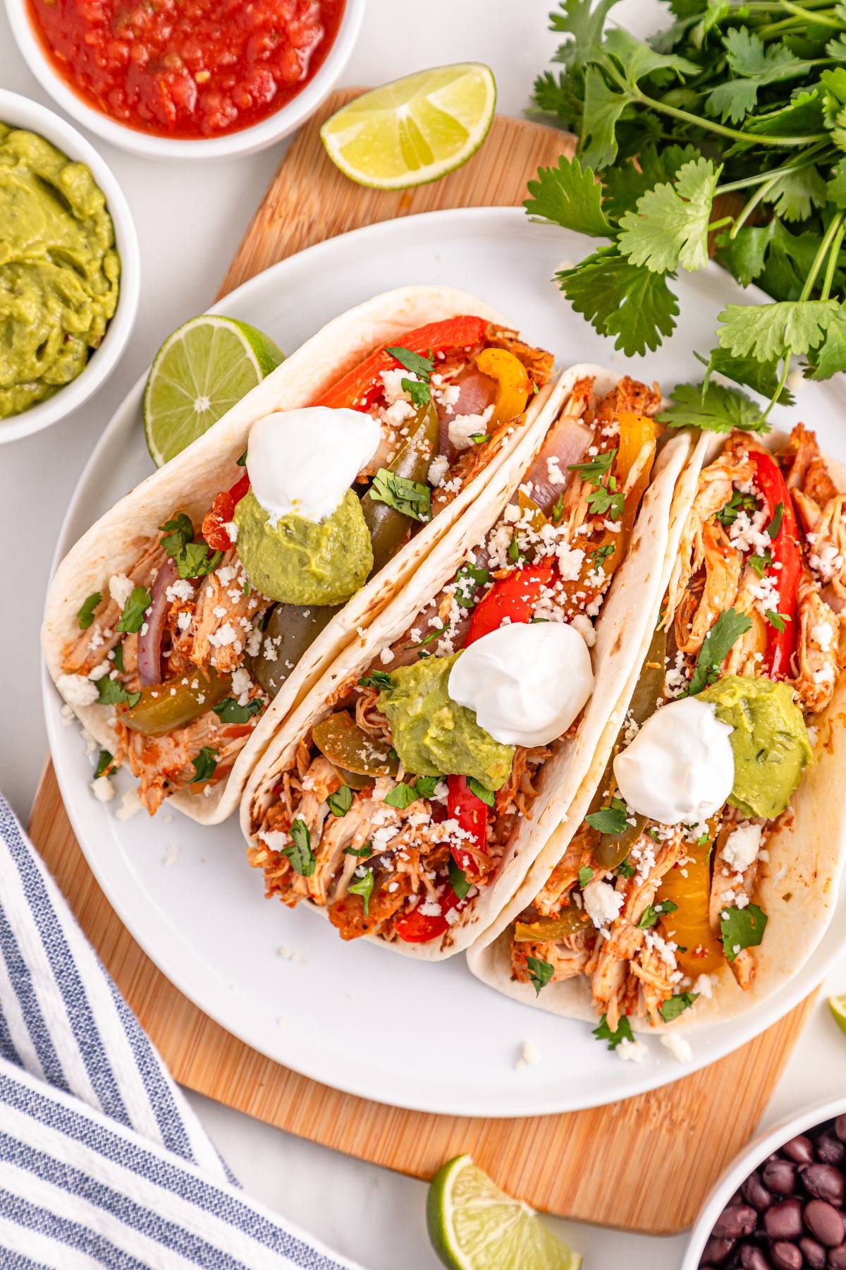 Three slow cooker chicken fajita topped with sour cream, guacamole, and cilantro on a white plate, with limes nearby.