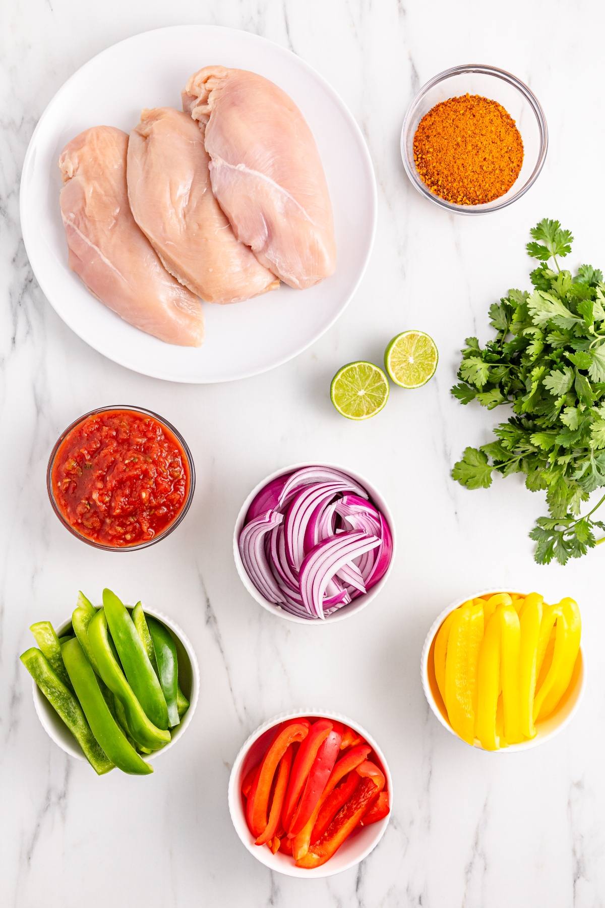 Raw chicken breasts on a plate with sliced peppers, onions, salsa, seasoning, cilantro, and lime on a marble surface.