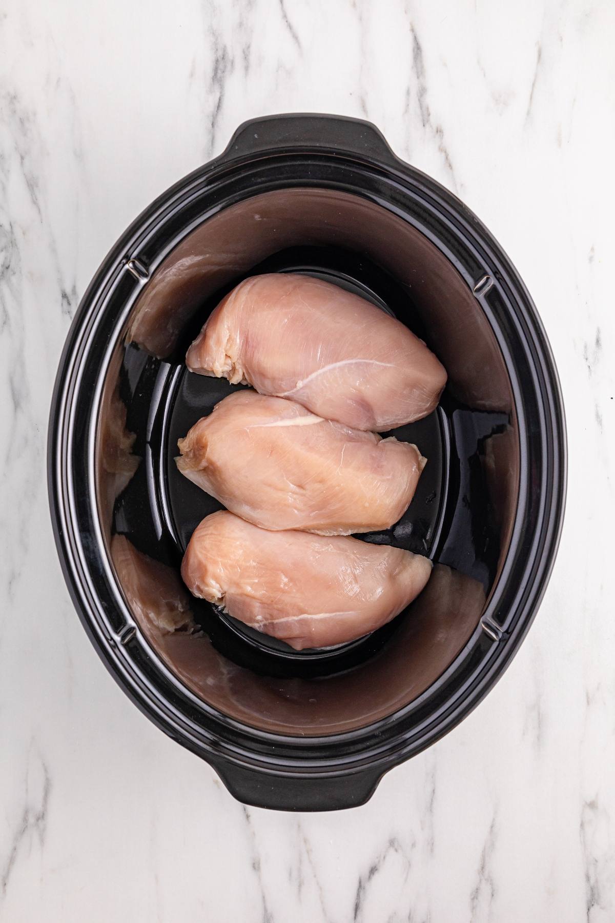 Three raw chicken breasts in a black slow cooker on a white marble surface.