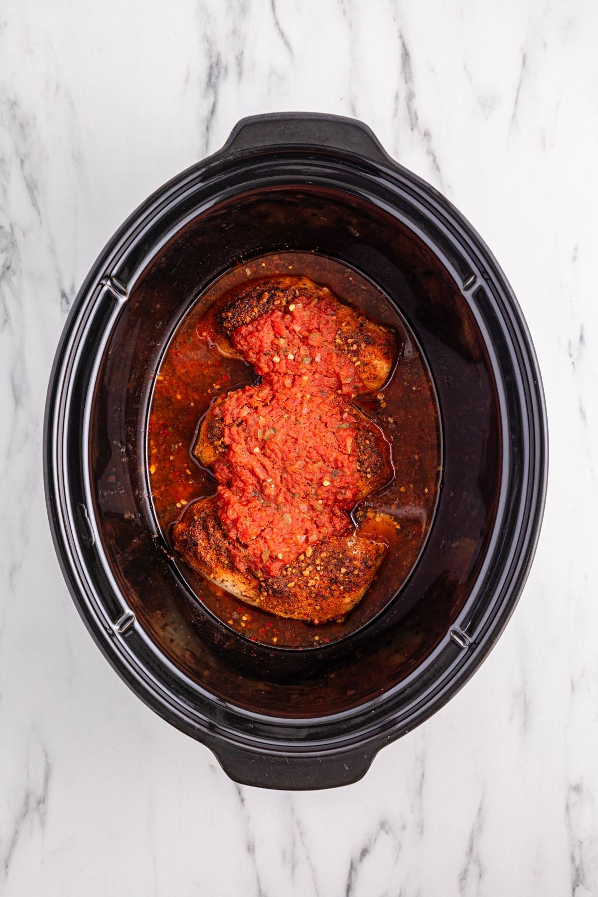 Raw seasoned meat topped with tomato sauce in a black slow cooker on a white marble surface.