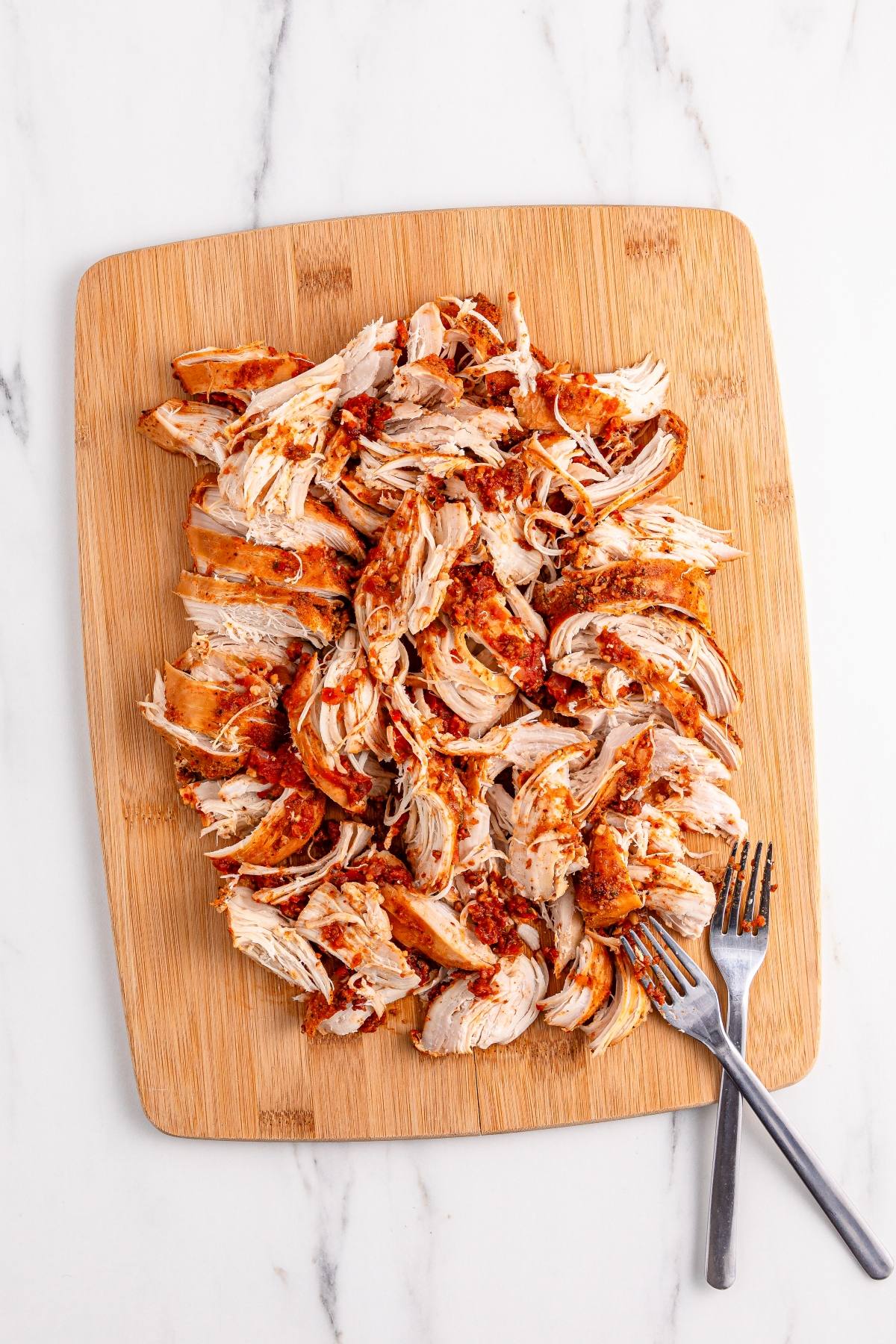 Shredded seasoned chicken on a wooden cutting board with two forks on the side.