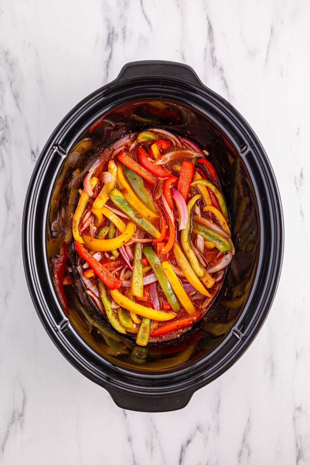 Sliced red, yellow, and green bell peppers with onions in a slow cooker on a white marble surface.