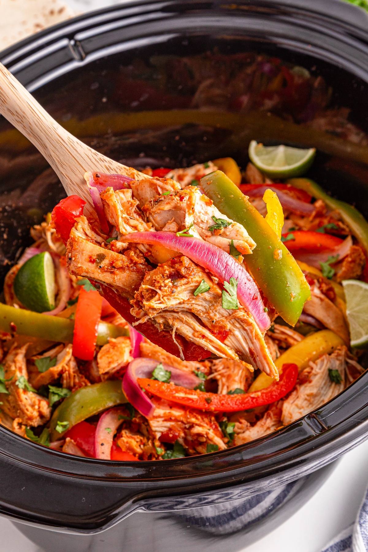A wooden spoon lifts cooked chicken, bell peppers, and onions from a slow cooker filled with fajita mix.