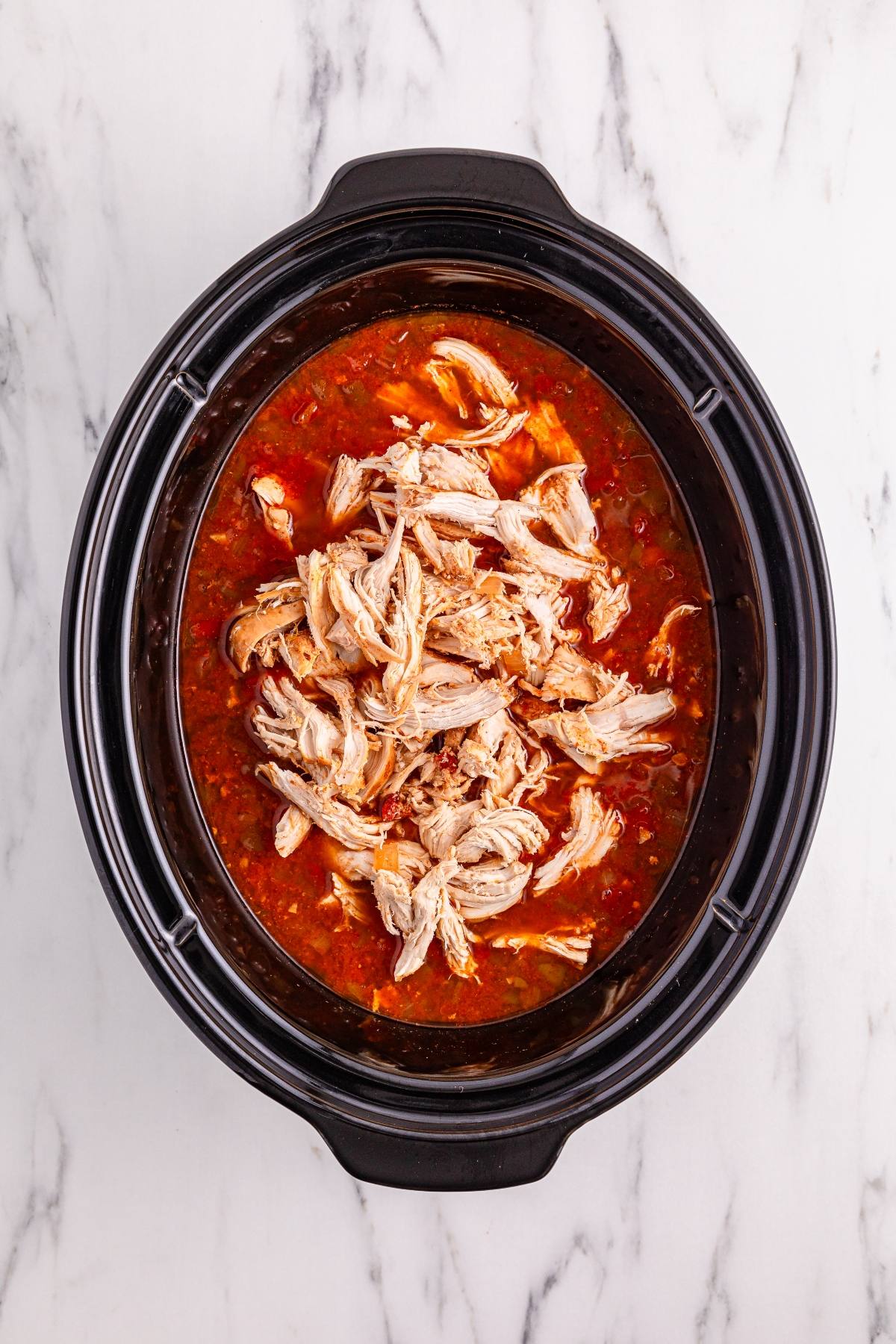 Shredded chicken in a rich red sauce inside a black slow cooker on a white marble surface.