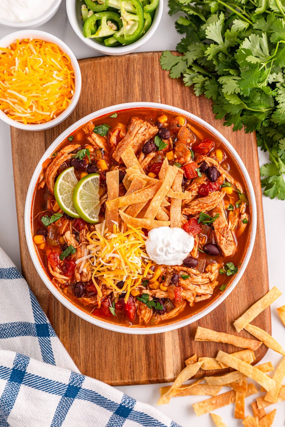 Bowl of chicken tortilla soup topped with cheese, sour cream, lime, and crispy tortilla strips, surrounded by toppings.