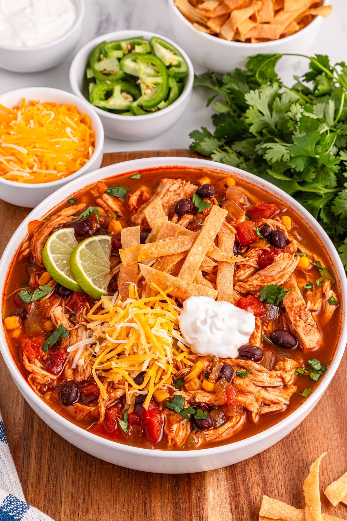 Bowl of chicken tortilla soup topped with shredded cheese, tortilla strips, lime, and sour cream, with toppings nearby.