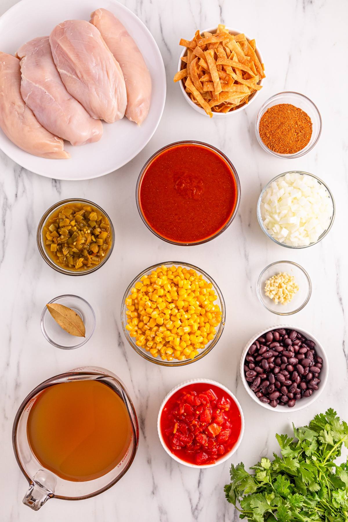 Ingredients for a chicken tortilla soup recipe arranged in bowls and plates on a marble countertop.
