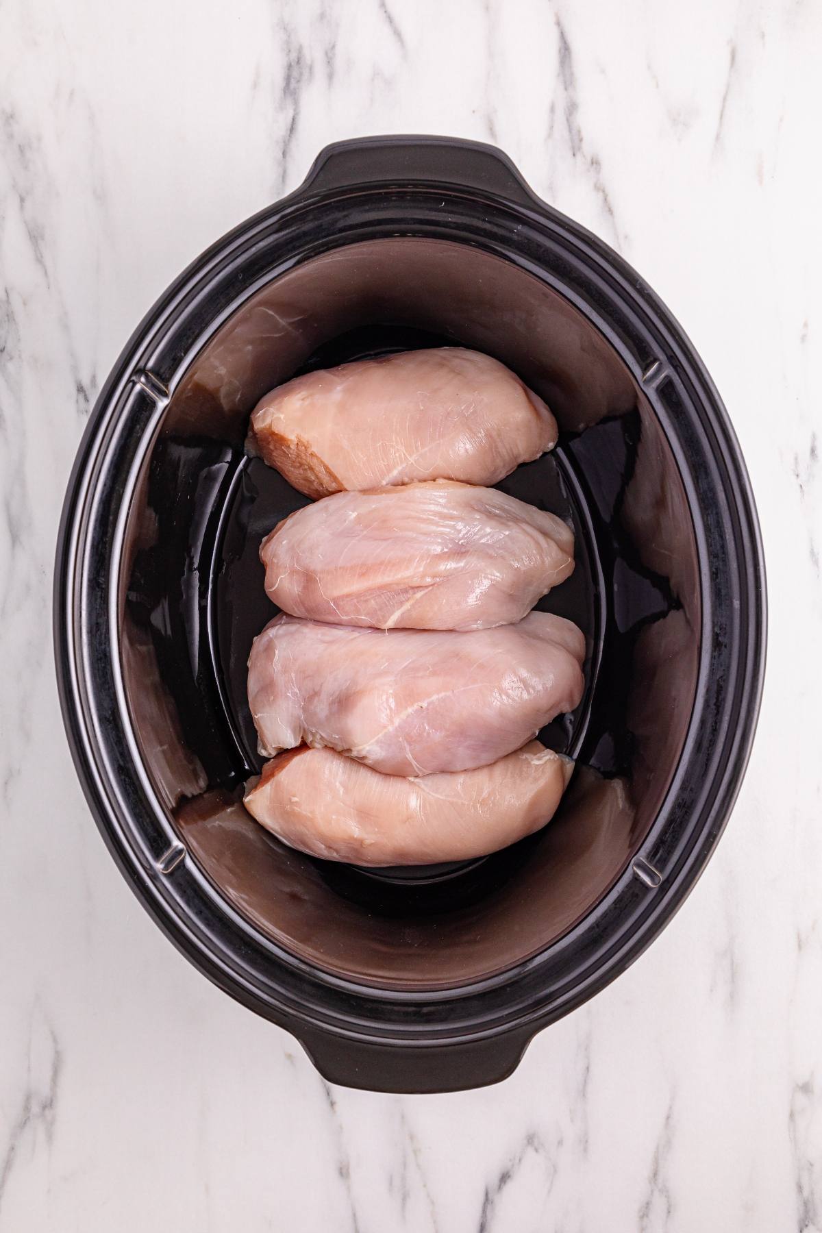 Four raw chicken breasts placed inside a black slow cooker on a white marble countertop.