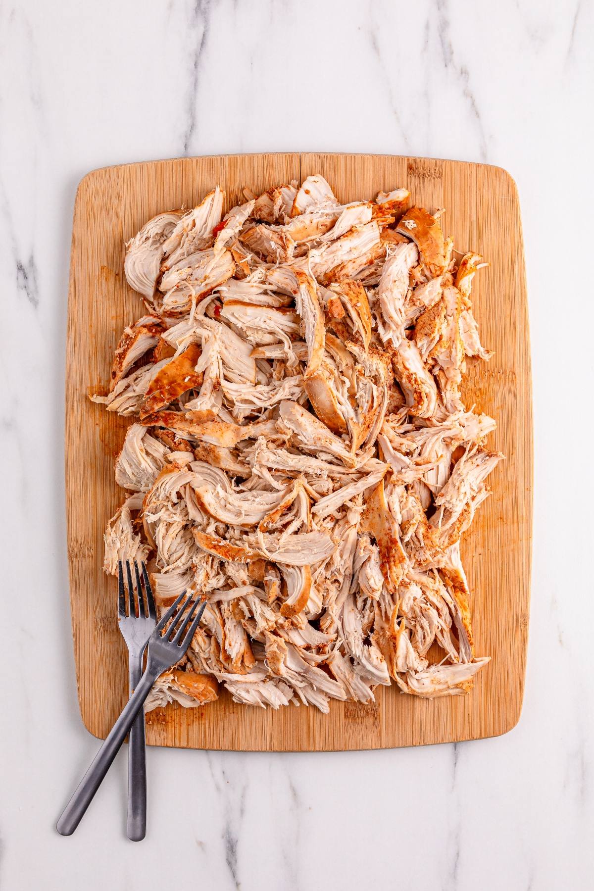 Shredded cooked chicken on a wooden cutting board with two forks beside it, on a marble surface.