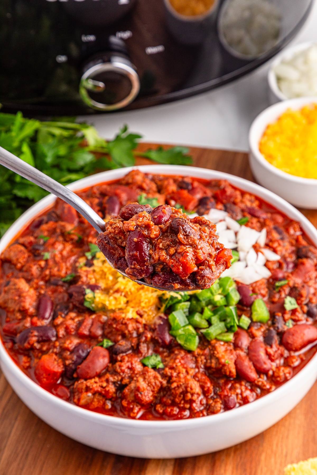 A spoonful of slow cooker chili with beans, meat, and tomatoes above a bowl topped with cheese, onions, and jalapeños.