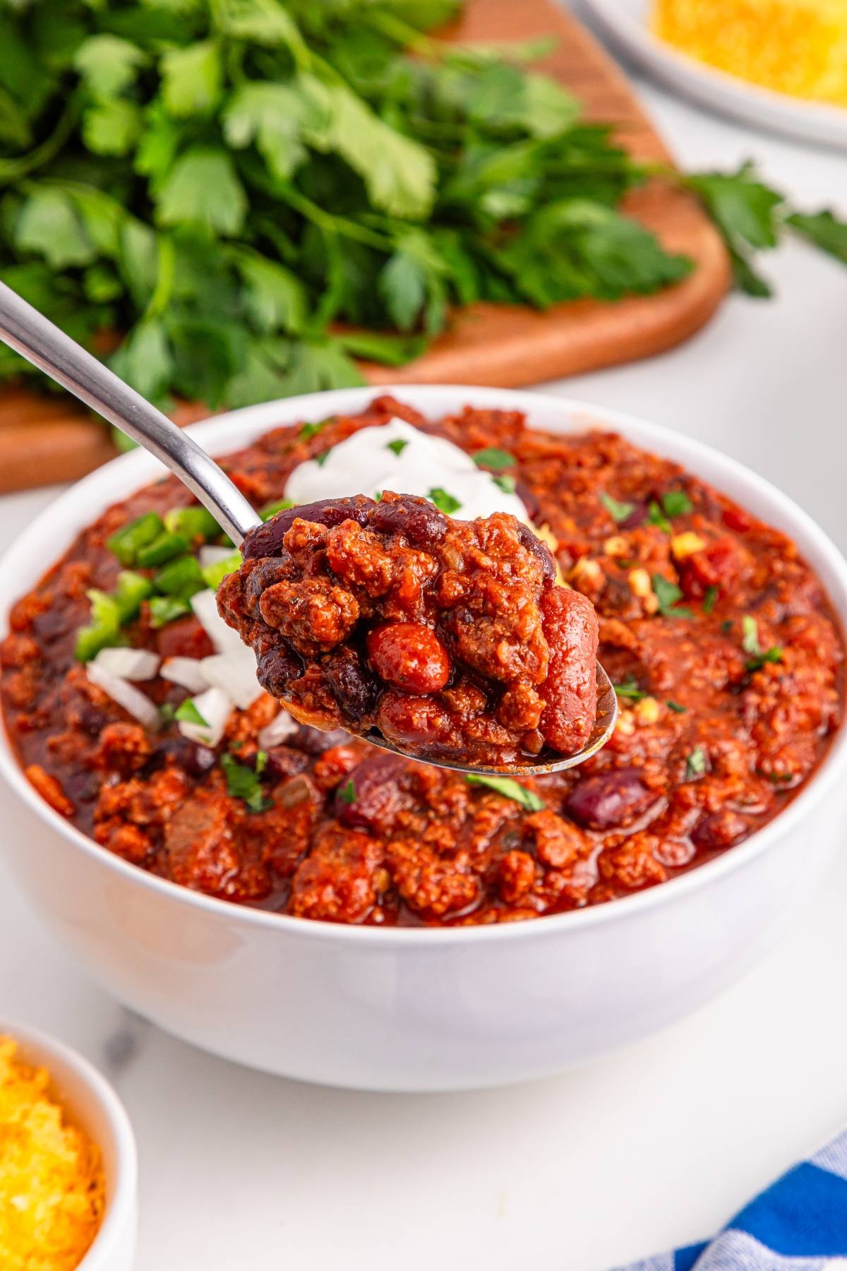 A spoonful of slow cooker chili with beans and ground meat, held above a bowl, topped with chopped onions and herbs.
