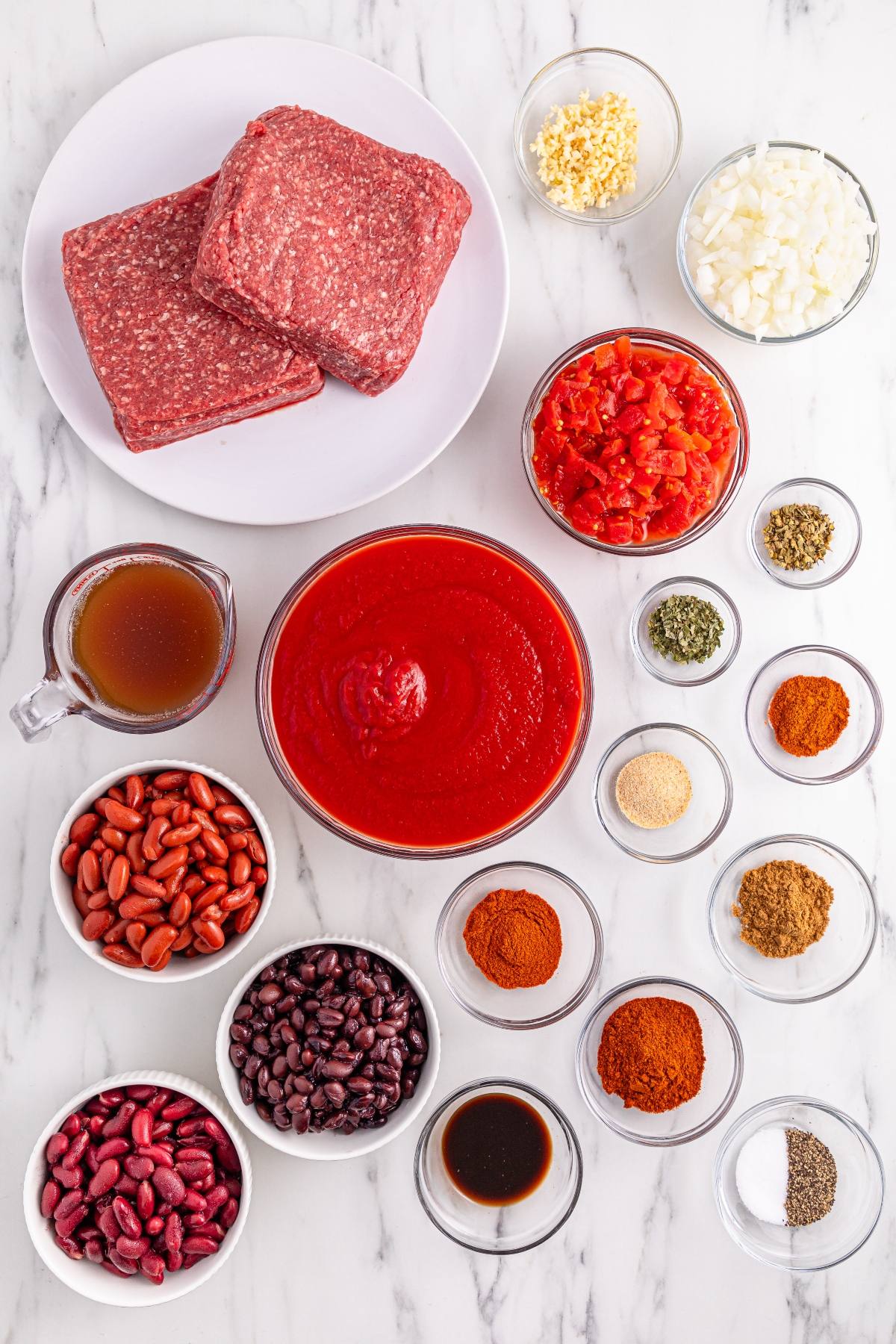 Ingredients for chili including ground beef, beans, tomatoes, spices, broth, onions, and garlic on a marble surface.