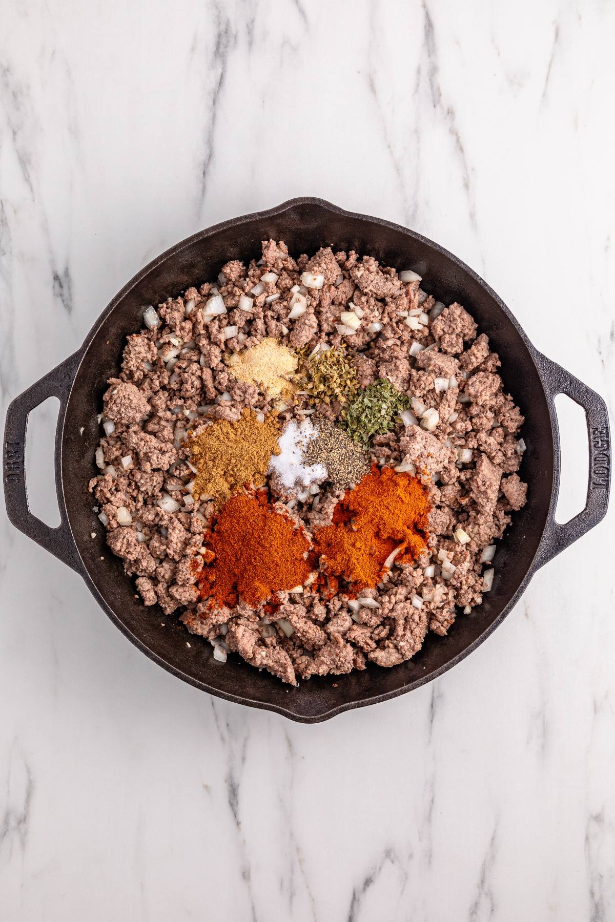 Ground beef and diced onions cooking in a skillet, topped with assorted spices on a marble countertop.