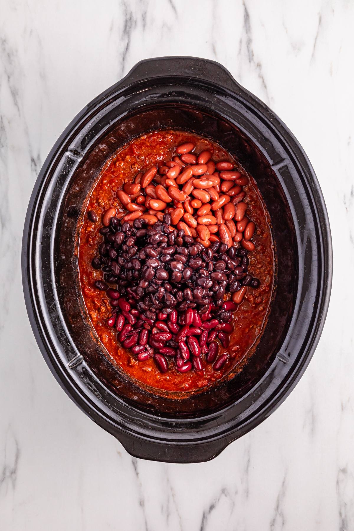Slow cooker with chili, topped with kidney beans, black beans, and pinto beans on a marble countertop.