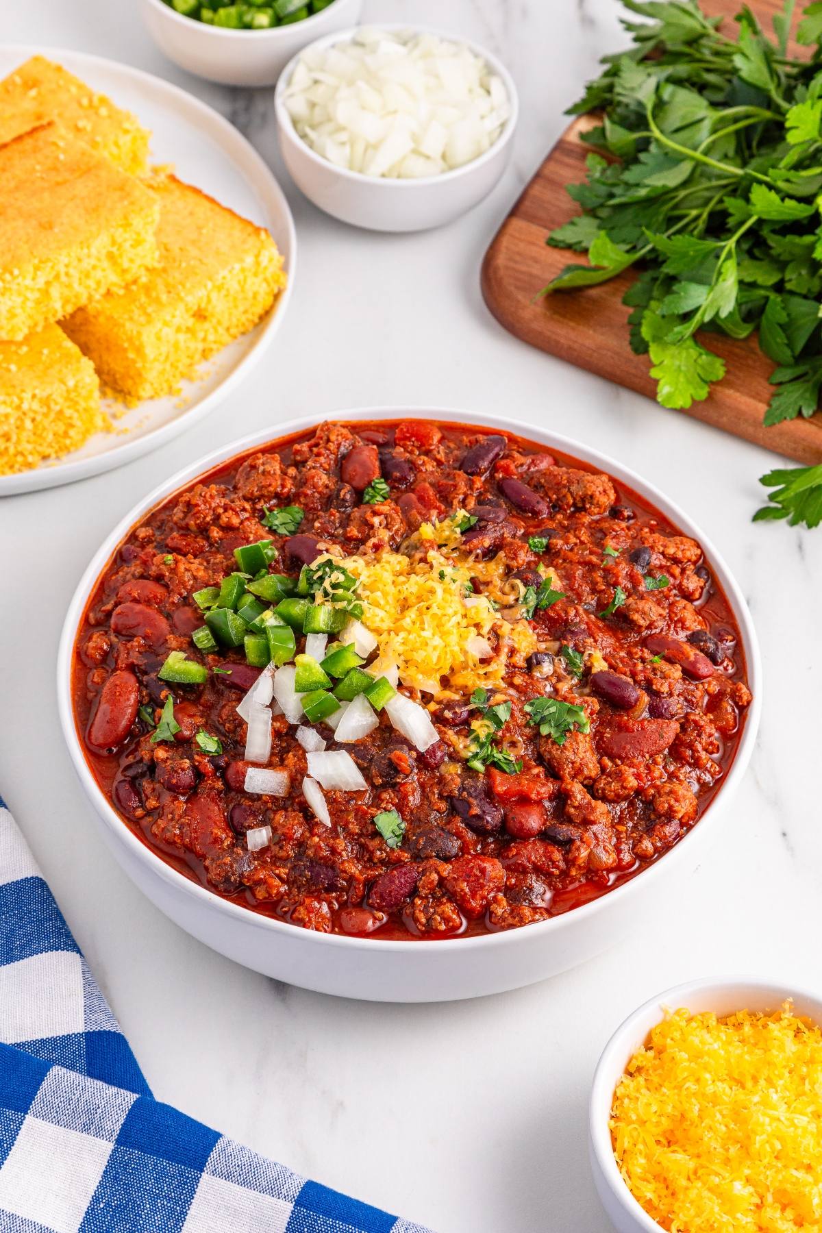 Bowl of slow cooker chili topped with cheese, onions, and jalapeños, with cornbread and fresh herbs on the side.