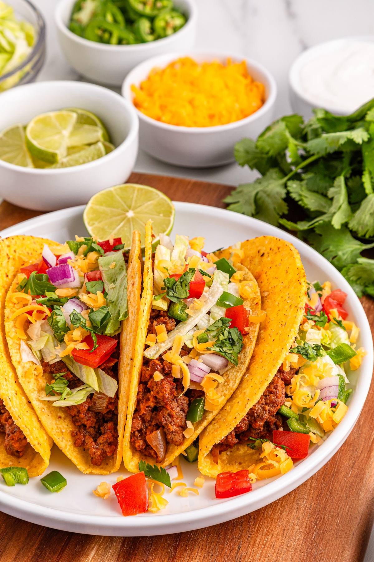 Three hard shell tacos filled with ground beef, lettuce, cheese, onions, and peppers on a white plate.