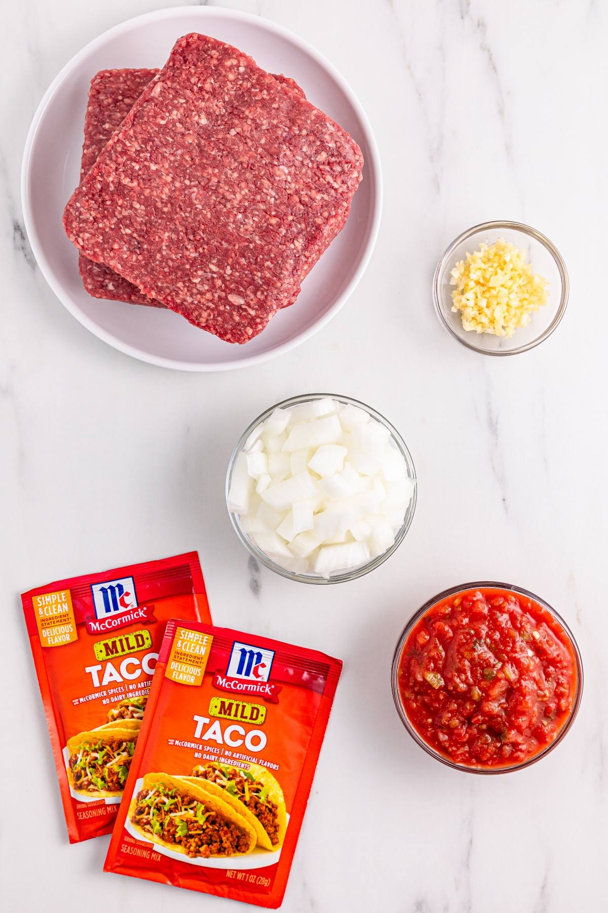 Ground beef, chopped onions, minced garlic, salsa, and taco seasoning packets arranged on a white surface.