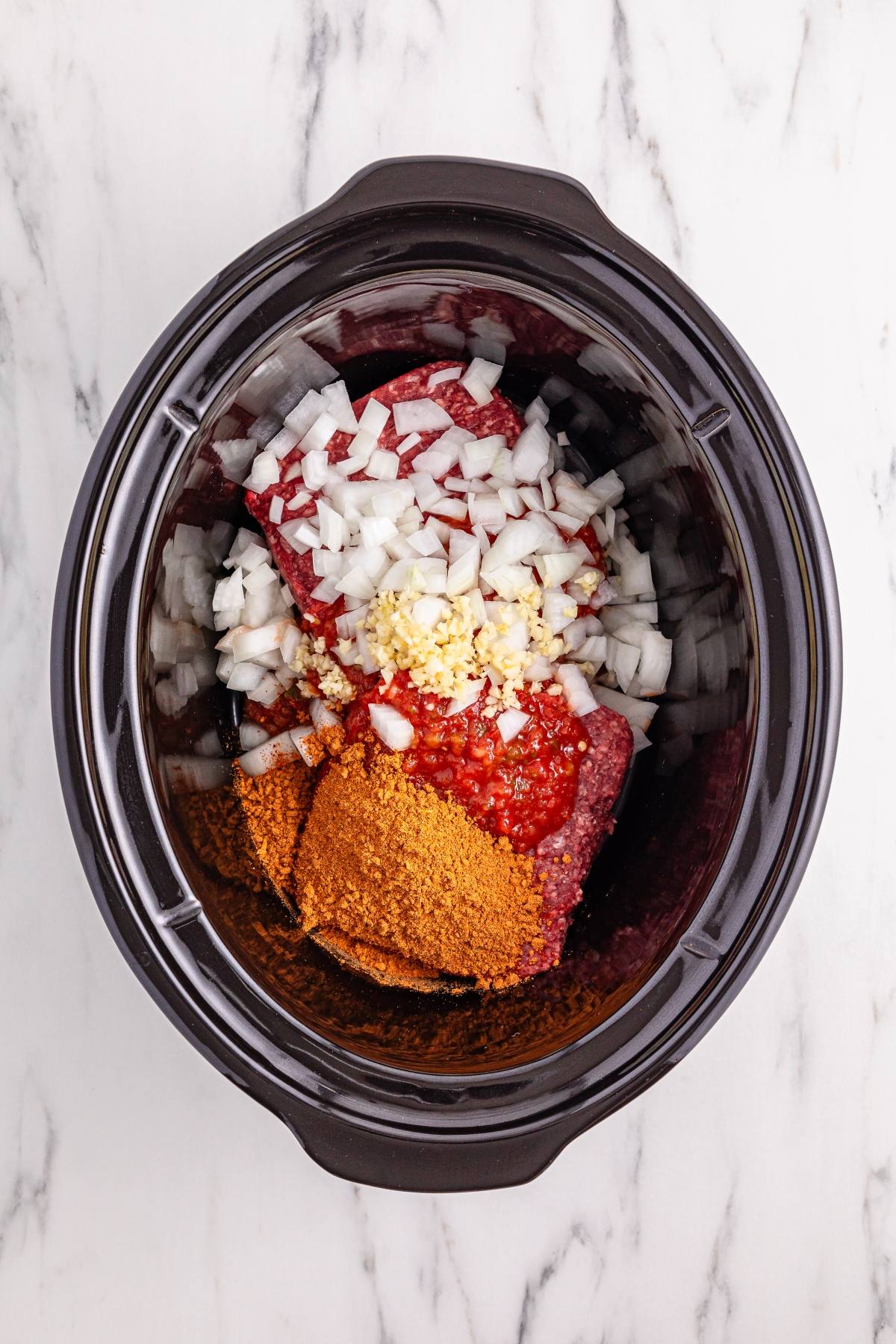 Chopped onions, ground meat, spices, tomatoes, and garlic in a slow cooker on a marble surface.