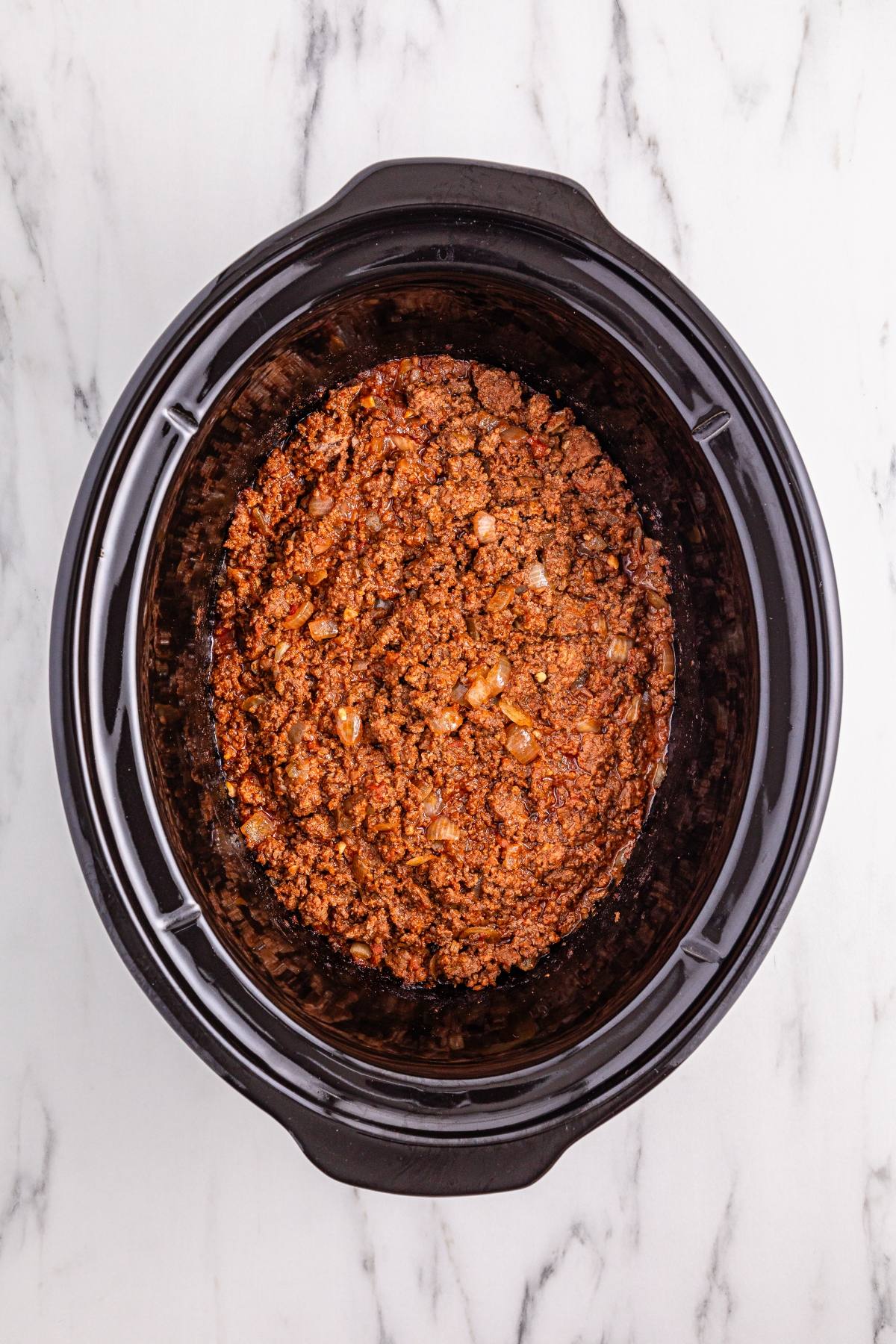 Ground beef cooked with spices in a black slow cooker on a white marble surface, viewed from above.