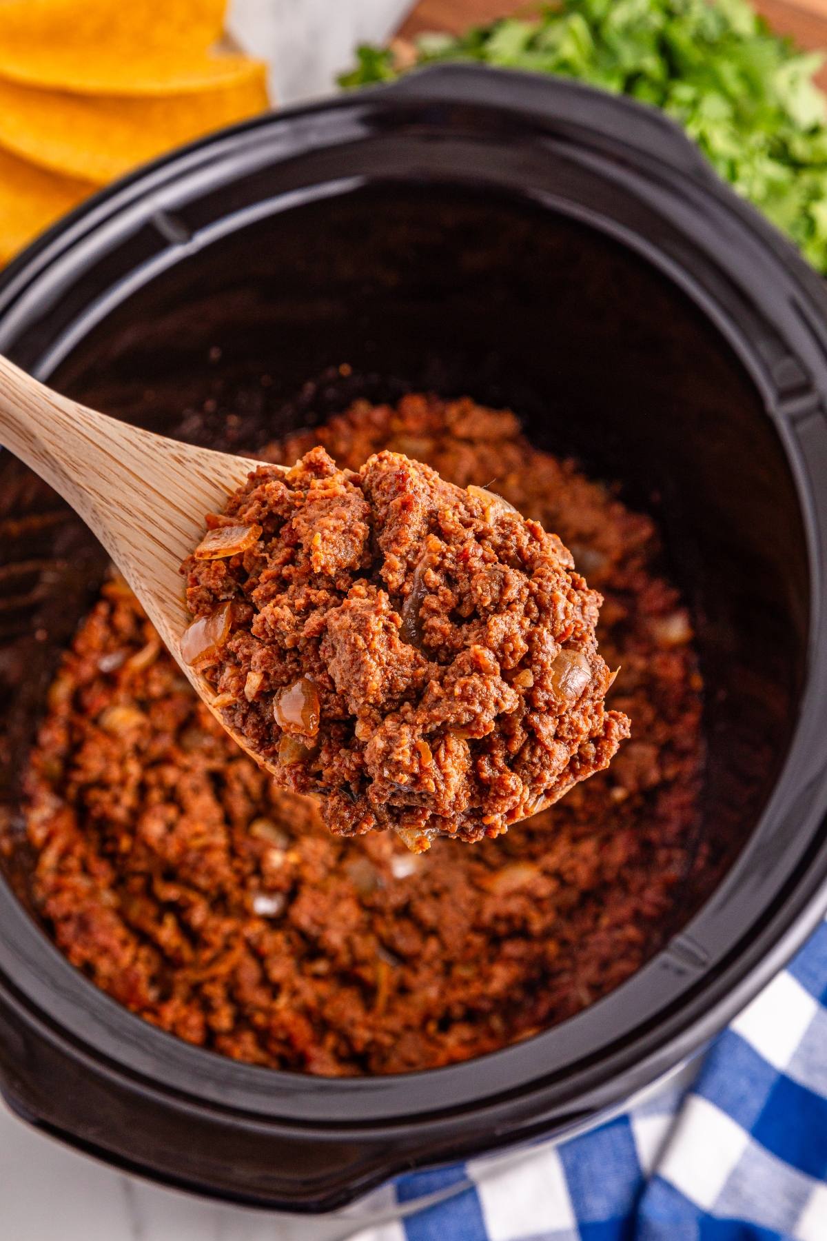 A wooden spoon holds cooked ground beef taco filling over a black slow cooker, with cilantro in the background.