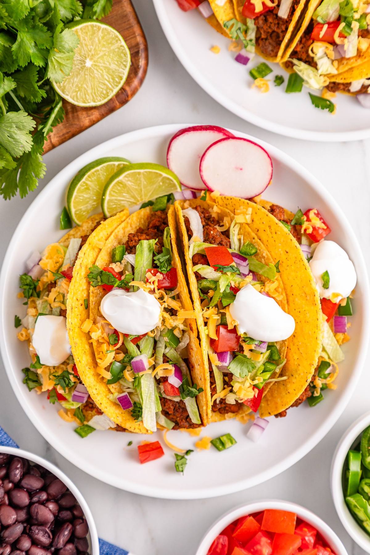 Three hard shell slow cooker tacos with ground beef, lettuce, cheese, veggies, and sour cream on a white plate with lime slices.
