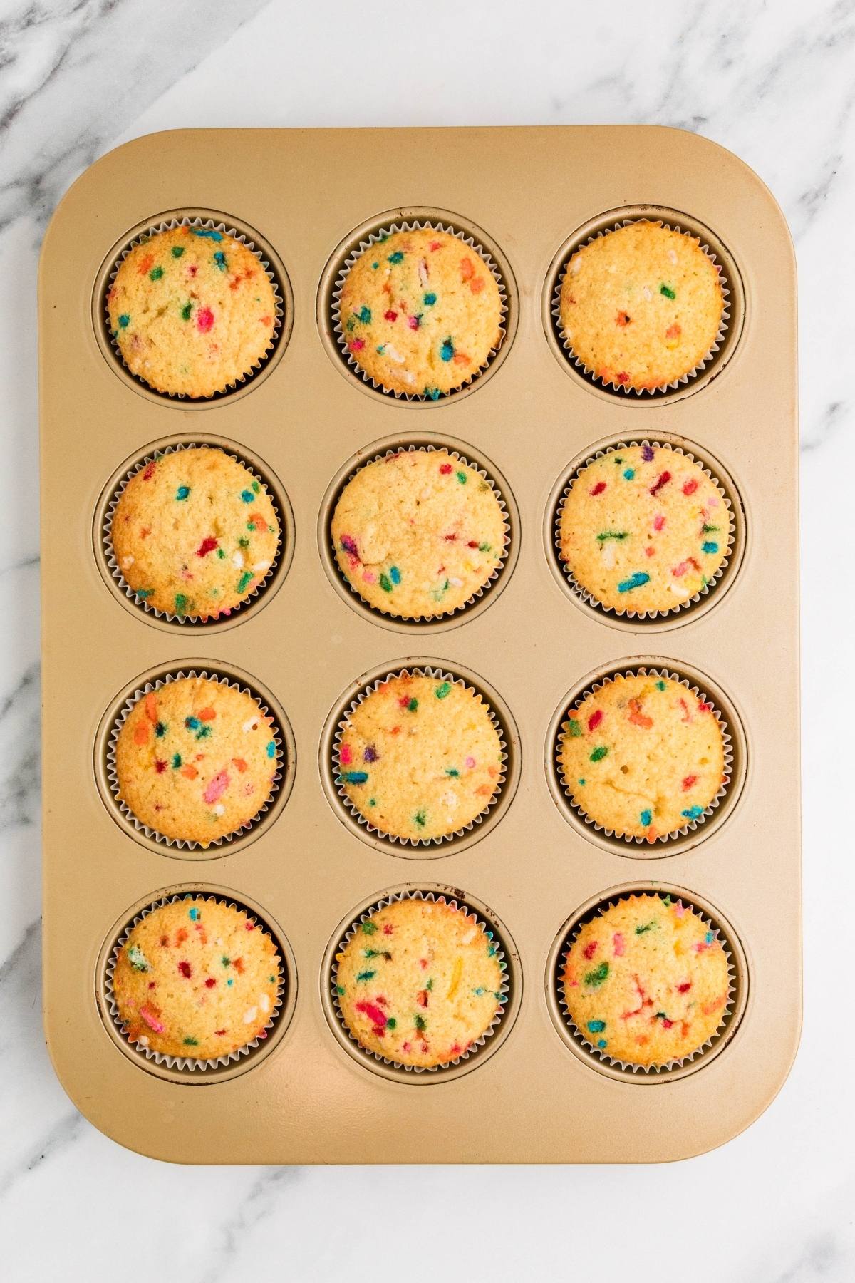 A muffin tray with 12 funfetti cupcakes in colorful paper liners on a marble surface.