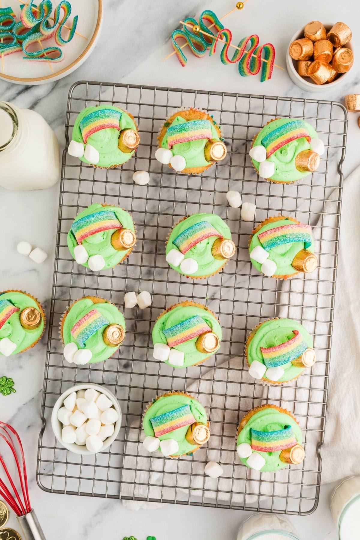 Cupcakes with green frosting, rainbow candy, marshmallows, and gold-wrapped chocolates on a cooling rack.