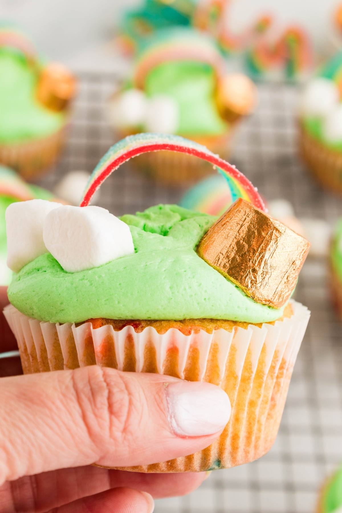 A hand holding a cupcake with green frosting, mini marshmallows, a gold-wrapped candy, and a rainbow candy strip.