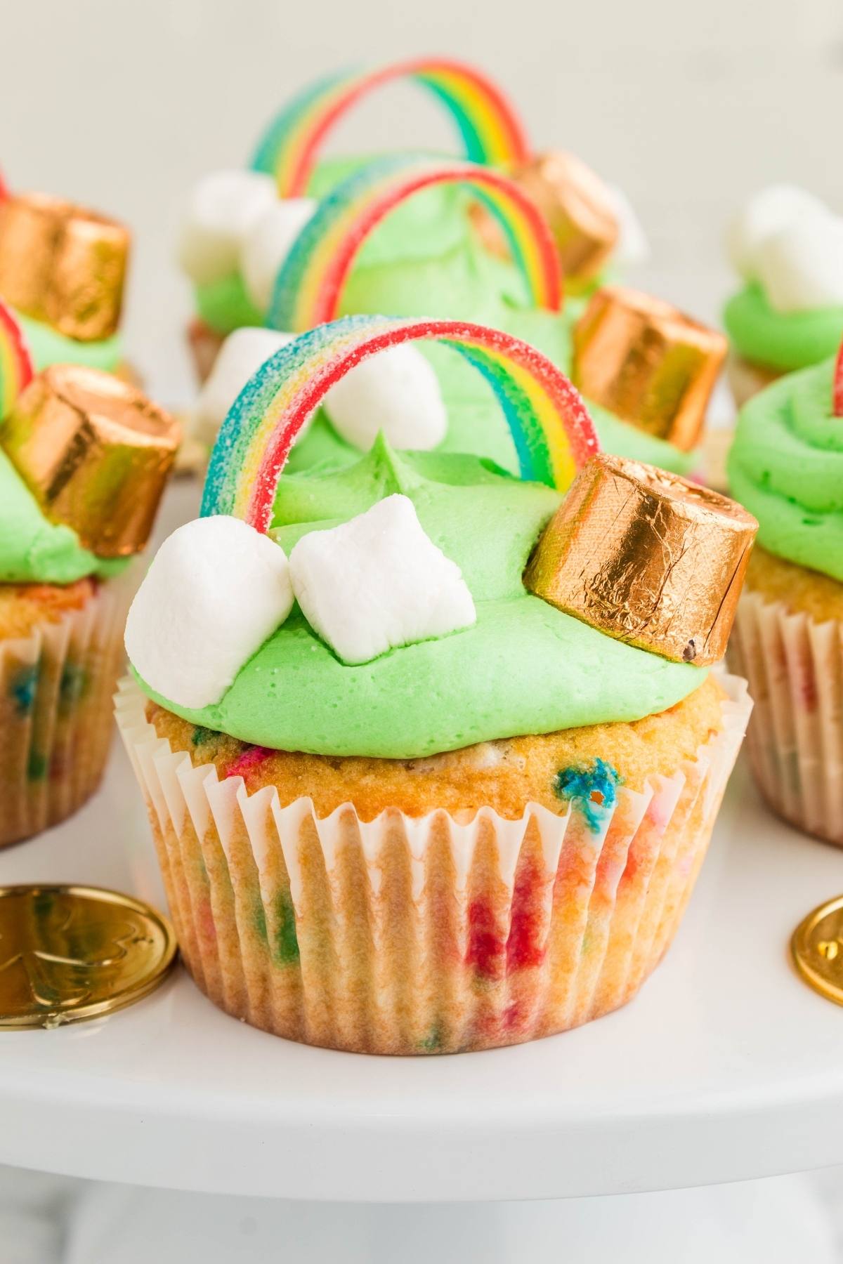 St. Patrick's Day Cupcake topped with green frosting, rainbow candy, gold chocolate, and mini marshmallows on a white stand.