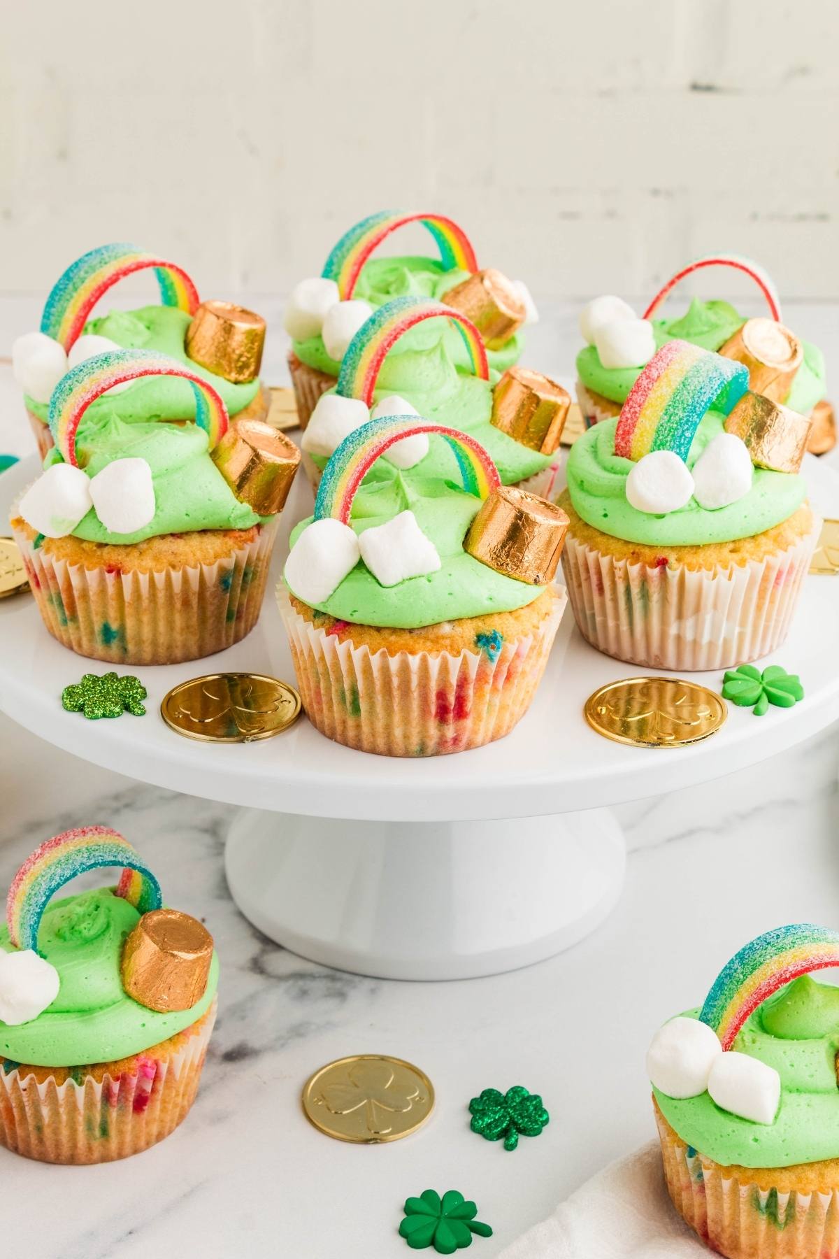 St. Patrick's Day cupcakes with green frosting, rainbows, gold coins, and marshmallows on a white stand.