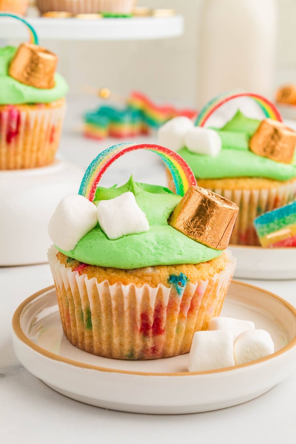 A St. Patrick's Day cupcake with green frosting, marshmallows, a gold-wrapped candy, and a rainbow candy strip on top.