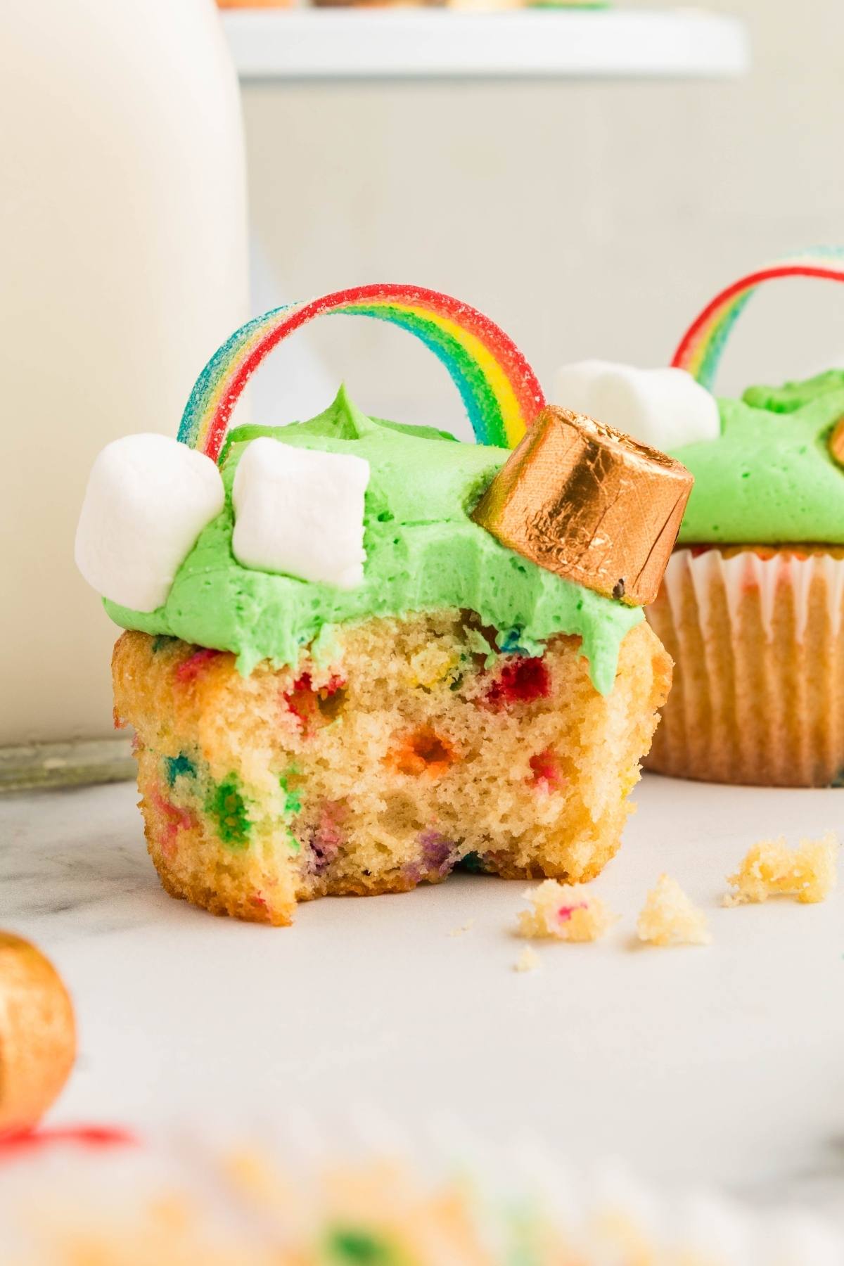 A St. Patrick's Day cupcake with green frosting, rainbow candy, marshmallows, and a gold-wrapped chocolate, with a bite taken out.