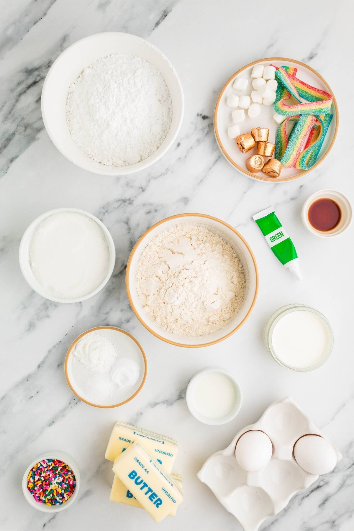 Baking ingredients on a marble surface, including flour, eggs, butter, milk, candies, and sprinkles.