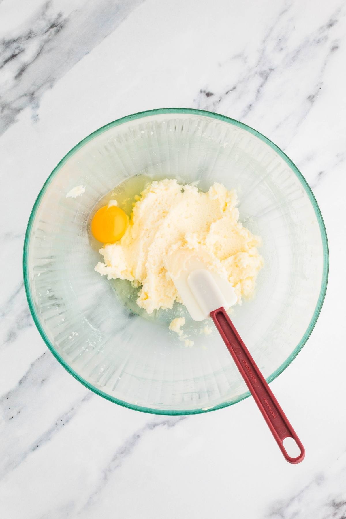 A mixing bowl with butter, sugar, and an egg, plus a spatula on a marble countertop.