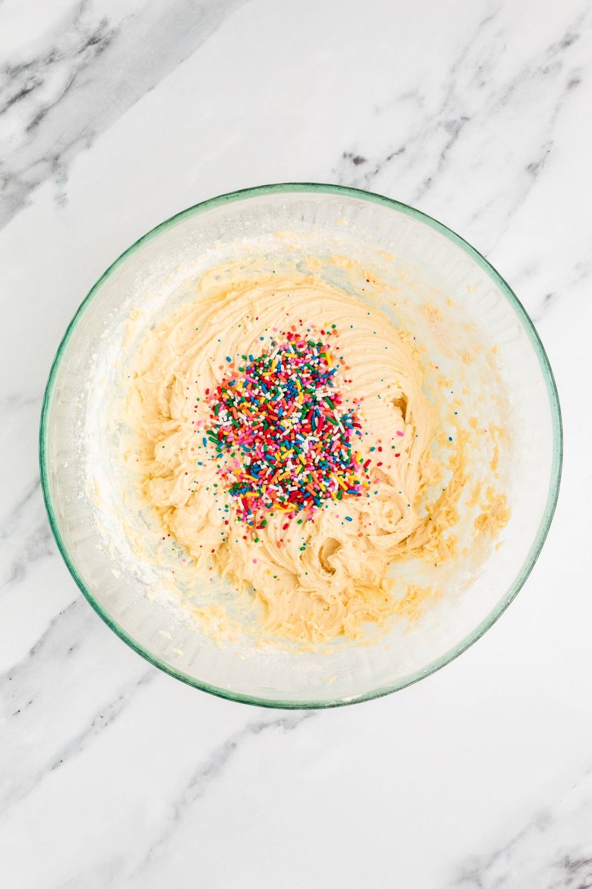Glass bowl with cookie dough topped with colorful rainbow sprinkles on a white marble surface.