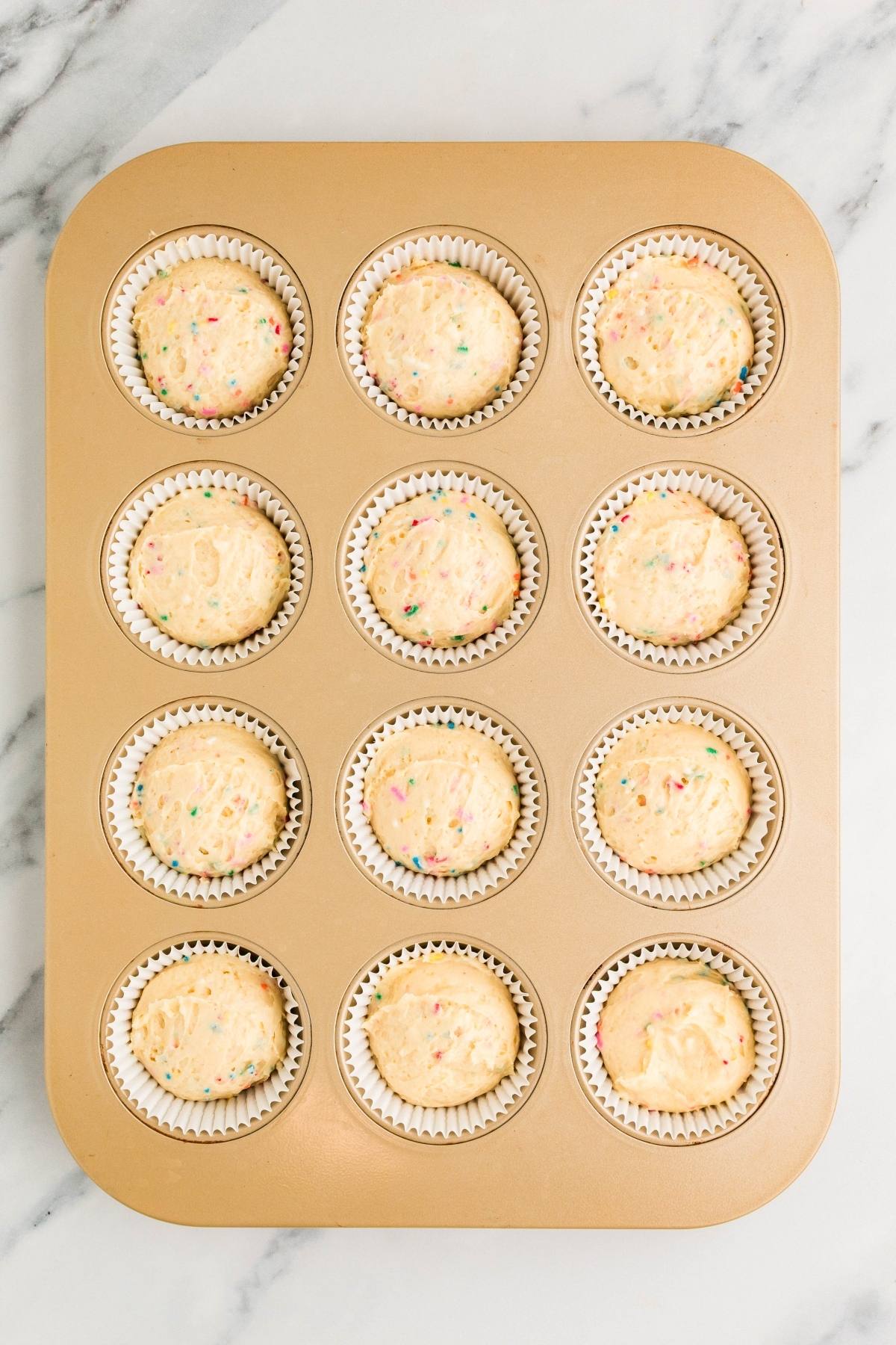 A gold muffin pan holds 12 cupcake liners filled with funfetti cupcake batter on a marble surface.
