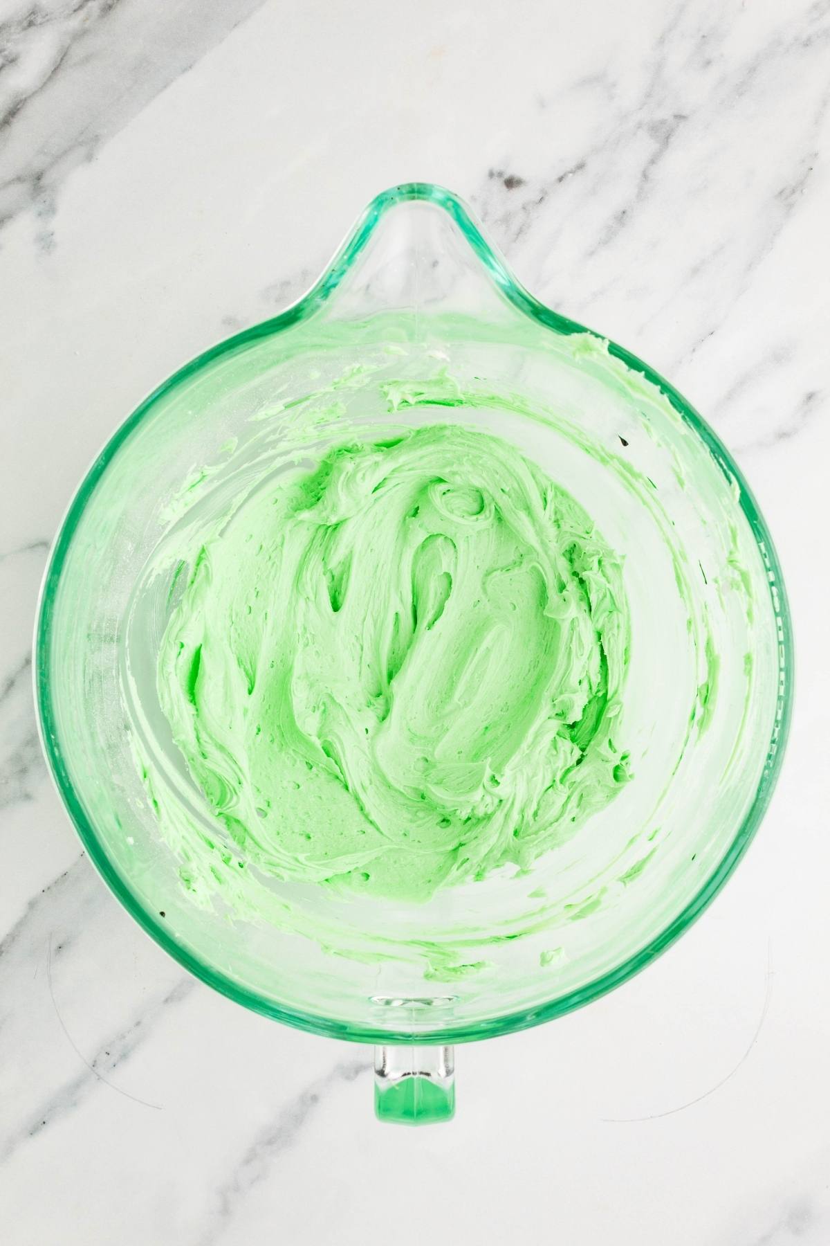 A glass mixing bowl with light green frosting on a white marble surface.
